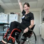 Ryann, a person with short brown hair and a black nursing outfit is sitting in a red wheelchair proudly holding her arm up high with her mouth open. She wears a stethoscope around her neck and several tattoos are visible on her arms.