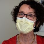 Dr. Wolf, a woman with short, curly brown hair, black glasses and a yellow face mask.