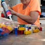 A person holding a big glue pistol sitting in a kitchen with a huge bag of colorful Lego in front.