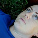 Caitlin, a woman with long red hair is lying in green grass. She wears a blue shirt.