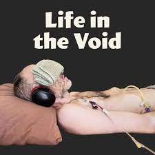 A photo of a man with tubes coming out of his upper body. He wears headphones and has his eyes covered. Text: Life in the Void