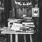 A black and white image of a small table near a wooden wall. The table is stacked haphazardly with books and papers.