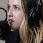 A close-up of a woman wearing headphones and speaking into a microphone. She has light brown, shoulder-length hair with blonde highlights. She looks forward with focus as she speaks into a black, professional studio microphone attached to a flexible arm. The background is blurred and neutral.