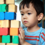 A child playing with colorful building blocks looking serious. The child has brown short hair and wears a blue shirt with red stripes.